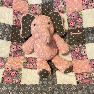 May include: A pink plush elephant toy with gray polka dot ears and feet sits on a patchwork quilt. The quilt features squares of pink floral fabric, white textured fabric, and gray floral fabric.