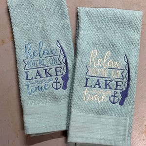 May include: Two light blue textured towels with embroidered text that reads "Relax, you're on lake time" with a fishing rod and anchor design. The towels are a light blue color.