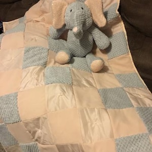 May include: A soft, plush elephant toy with light blue fur, beige ears, and feet sits on a patchwork baby blanket. The blanket features alternating squares of light blue textured fabric and beige satin.
