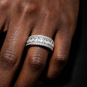 May include: A wide silver-toned band ring featuring a central row of large, round, clear stones flanked by two thinner rows of smaller, pavé-set clear stones. The ring is worn on the ring finger, showcasing a metallic finish and reflective surfaces.