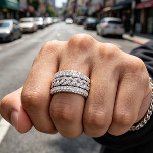 Pode incluir: Um anel largo de tom prateado com três bandas distintas. As bandas superior e inferior estão incrustadas com pequenas pedras claras e brilhantes, enquanto a banda central apresenta um design texturizado de elos de corrente entrelaçados.