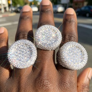 May include: Three silver-toned rings featuring large, circular faces completely encrusted with numerous small, clear, sparkling stones. The rings are worn on three fingers, showcasing a dense, pavé-style setting that covers the entire surface of each ring.