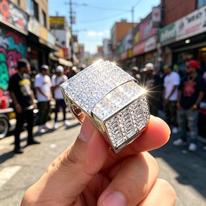 May include: A large silver-toned ring featuring a wide, rectangular face completely covered in numerous clear, sparkling stones arranged in a grid pattern, with additional rows of baguette-cut stones along the sides, reflecting bright sunlight.