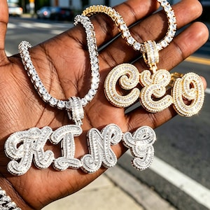 Op de afbeelding: Twee metalen kettingen met tennis-stijl schakels en heldere stenen. De ene ketting is zilverkleurig met een hanger in cursief schrift die KING spelt, de andere is goudkleurig met een hanger die CEO spelt in cursief schrift.