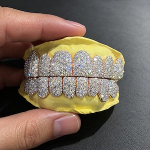 Peut inclure: Un ensemble de grillz dentaires dorés comprenant deux rangées de dents recouvertes de nombreuses petites pierres claires et scintillantes. Les grillz sont montés sur un moule dentaire jaune, présentant un sertissage dense de style pavé.