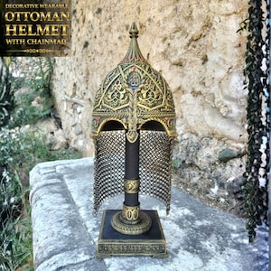 Peut inclure: Un casque ottoman décoratif avec cotte de mailles, en métal doré avec des motifs en relief complexes, un sommet pointu et de petites pierres rouges et bleues incrustées. Le casque est présenté sur un socle assorti doré et noir.