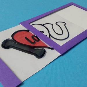 May include: A rectangular white card with a purple frame featuring a sliding mechanism. The card displays a black letter I, a red heart, and the letters L, O, V, and E in black outline. The sliding purple frame reveals the text I Love U.