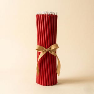 May include: A bundle of red taper candles tied with a gold ribbon. The candles have white wicks and are arranged in a vertical stack. The ribbon is tied in a bow around the center of the candles. The background is a solid, light beige color.