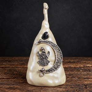 May include: A cone-shaped cream-coloured sculpture with a smooth, pearlescent finish. It features a small black stone at the top, a silver-toned metal crescent moon with intricate filigree patterns, and a silver-toned metal angel charm.