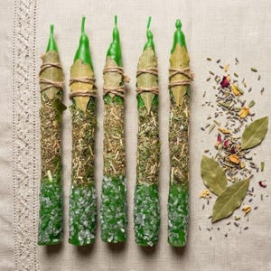 May include: Five green spell candles are arranged on a neutral-toned surface. Each candle is adorned with dried herbs, bay leaves, and twine. The base of each candle is covered in small, white crystals. Dried herbs and bay leaves are scattered nearby.