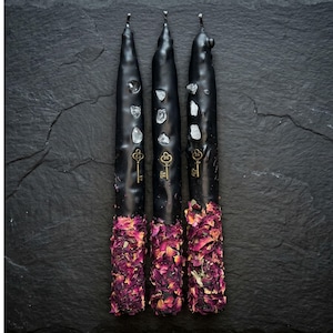 May include: Three black taper candles with embedded crystals and antique key charms. The base of each candle is adorned with dried rose petals. The candles are displayed on a dark, textured surface.