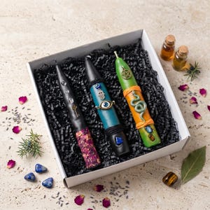 Op de afbeelding: Een witte geschenkdoos met drie taps toelopende kaarsen in zwarte papieren snippers. Eén kaars is zwart met gedroogde rozenblaadjes en een gouden sleutelbedel. De tweede is blauw met een hamsa- en oogbedel. De derde is groen en oranje met een slang en een pentagram.