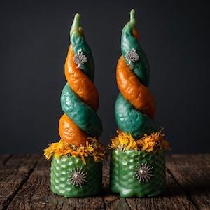 May include: Two spiral-shaped candles featuring alternating orange and green wax segments. Each candle has a silver tree charm near the top and a green honeycomb-textured base adorned with dried yellow flower petals and a silver sun charm.