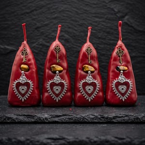May include: Four red candles in a teardrop shape, each decorated with a silver heart pendant with an eye design. A key charm and a tiger's eye stone are also attached. The candles are displayed on a dark surface.