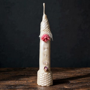 Op de afbeelding: Een lange, gebroken witte taps toelopende kaars met een getextureerde basis, versierd met een stuk wit kanten net bij de top, een kleine roze bloem, drie ingebedde heldere kristallen en een enkele gedroogde rozenknop onderaan de kaars.