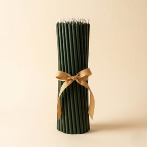 Puede incluir: Un paquete de velas cónicas verde oscuro atadas con una cinta dorada. Las velas tienen mechas blancas y están dispuestas verticalmente. La cinta está atada con un lazo alrededor del centro de las velas. El fondo es beige claro.