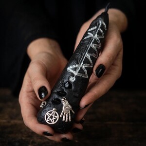 May include: A black textured candle featuring a silver hand-painted runic symbol, a silver pentagram charm, a silver hamsa hand charm, and a small silver skull charm embedded into the wax surface. The candle has a tapered shape and a dark finish.