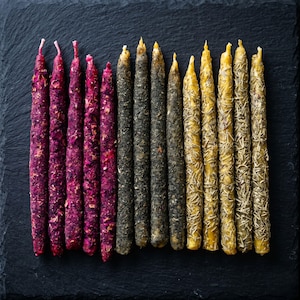 Puede incluir: Una variedad de velas decorativas delgadas en fila. Las velas son de color rojo oscuro, negro y amarillo. Algunas están adornadas con pétalos o hierbas secas, agregando textura. Las velas miden unos 20 cm de largo y están sobre una superficie oscura.