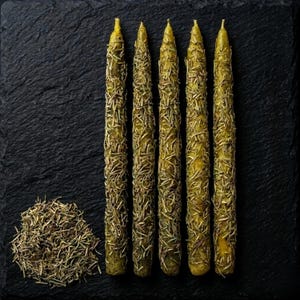 May include: Five green taper candles, adorned with dried rosemary, are arranged on a dark, textured surface. A small pile of loose rosemary sits to the left of the candles. The candles have a rustic, natural appearance.