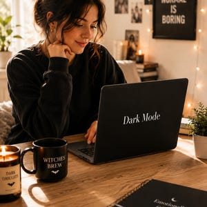 May include: A black laptop displaying "Dark Mode" on a wooden desk, next to a black mug with "Witches Brew" and a candle labelled "Dark Thoughts". A notebook with "Emotionally Offline" is also on the desk, creating a dark aesthetic.