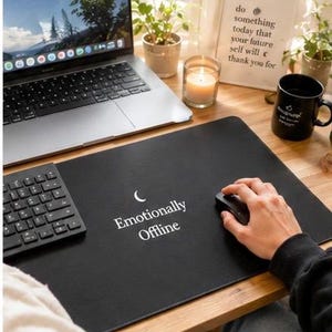 Könnte beinhalten: Schwarze rechteckige Schreibtischunterlage mit dem weißen Schriftzug "Emotionally Offline" und einem Halbmond-Symbol. Eine Hand bedient eine Maus auf der Unterlage. Ein Laptop, eine Tastatur und eine Tasse sind ebenfalls zu sehen.