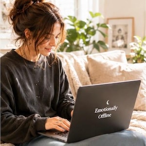 Op de afbeelding: Een zwarte laptop met de witte tekst "Emotionally Offline" is open op de schoot van een persoon. De persoon draagt een donkergrijze trui en zit op een bank. De achtergrond omvat een plant en een ingelijste foto.