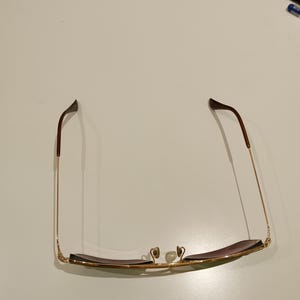 以下が含まれることがあります： ゴールドのメタルフレームと茶色のレンズのサングラス。テンプルは細く金色で、茶色のイヤーピースが付いています。ノーズパッドはクリアです。サングラスは白い面に置かれています。