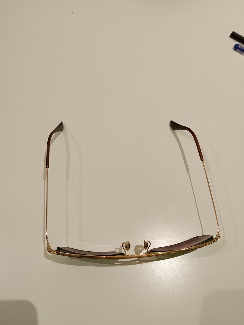 以下が含まれることがあります： ゴールドのメタルフレームと茶色のレンズのサングラス。テンプルは細く金色で、茶色のイヤーピースが付いています。ノーズパッドはクリアです。サングラスは白い面に置かれています。