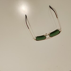 以下が含まれることがあります： 緑色のレンズが付いた金色のサングラス。 サングラスはダブルブリッジと、茶色の先端が付いた細いテンプルを備えています。 レンズは長方形で、フレームは明るい金色です。 サングラスは白い表面に置かれています。