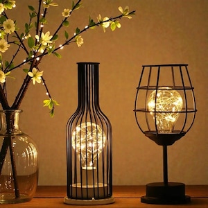 Op de afbeelding: Decoratieve zwarte metalen lampen in de vorm van een wijnfles en een wijnglas. Elke lamp heeft een warm wit LED-licht. Links van de lampen staat een glazen vaas met een bloeiende tak.