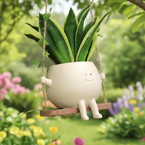 Puede incluir: Una maceta de cerámica de color crema en forma de personaje sonriente, suspendida de una rama de árbol con una cuerda. Contiene una planta verde vibrante con hojas bordeadas de amarillo. La maceta tiene brazos y piernas, y está sentada en un pequeño columpio de madera.