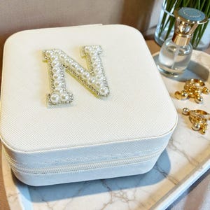 May include: A white jewelry box with a pearl and rhinestone letter "N" on the top. The box has a zipper closure and a textured surface. Other jewelry and a perfume bottle are in the background.