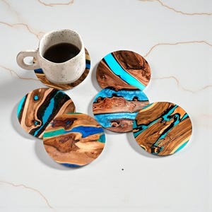 Può includere: Una tazza bianca macchiata con liquido scuro poggia su un sottobicchiere di legno. Diversi sottobicchieri rotondi con un motivo a venature del legno e accenti di resina blu sono sparsi. I sottobicchieri sono in legno e resina.