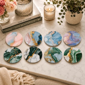 Puede incluir: Un juego de ocho posavasos circulares con diversos patrones de mármol en tonos rosa, azul, verde y morado, acentuados con vetas de color dorado. Cada posavasos muestra un diseño fluido único, dispuesto en dos filas sobre una superficie blanca.