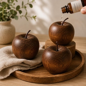 Op de afbeelding: Drie donkerbruine houten appelvormige ornamenten met zichtbare houtnerf en dunne steeltjes. Een klein amberkleurig glazen flesje met het label Lavender Essential Oil wordt boven een appel gehouden, waarbij een druppel olie naar beneden valt.