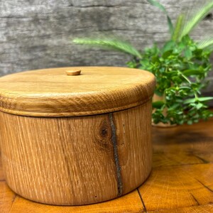 Op de afbeelding: Een cilindervormige houten container met een bijpassend rond deksel. De container is gemaakt van lichtbruin eikenhout, met een opvallend natuurlijk houtnerfpatroon en een zichtbaar verticaal knoestdetail aan de zijkant van de behuizing.