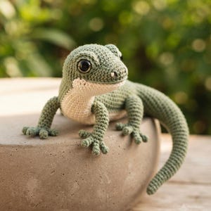 Peut inclure: Une figurine de lézard crochetée présentant un corps vert sauge avec un ventre crème, de grands yeux en plastique noir et quatre pattes détaillées avec des doigts. La créature rembourrée a une longue queue courbée et une surface en fil texturé.