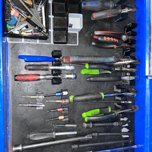 May include: A blue tool chest drawer containing an assortment of hand tools organized in black plastic holders. The collection includes various ratchets, pliers with red and green handles, screwdriver bits, extensions, and a bin of small hardware.