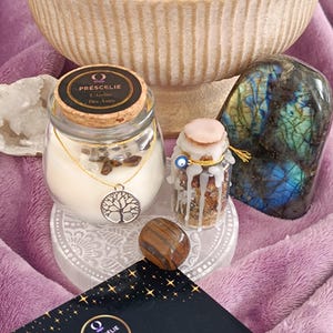 Peut inclure: Une collection d'articles spirituels, dont une bougie dans un bocal en verre avec un pendentif arbre de vie, un petit bocal avec un couvercle rose, une pierre précieuse polie et une pierre de labradorite. Une brochure avec le texte "PRÉSCÉLIE L'Atelier Des Ames" est également présente.