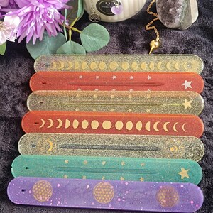 May include: Seven rectangular resin incense holders arranged in a stack. The holders feature various colors including clear with gold moon phases, red with stars, gold glitter, orange with moon phases, black glitter, teal with stars, and purple with gold geometric patterns.