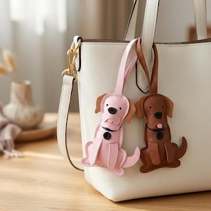May include: A white textured leather tote bag with two dog-shaped bag charms hanging from the handle. One charm is light pink with a black nose and collar, and the other is brown with a black nose, collar, and a small pink tongue detail.