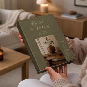 May include: A hardcover book with a matte olive green cover featuring the white text Rooted in Nature, Calm Spaces, Real Living. The cover displays a central photograph of a brown ceramic vase on a wooden table and a white line drawing of foliage.