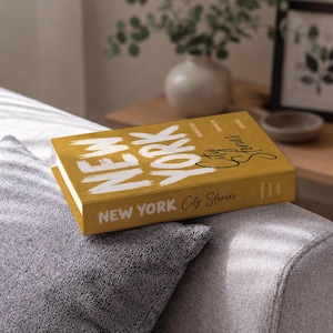 Könnte beinhalten: Ein senfgelbes Hardcover-Buch mit dem Titel New York City Stories, das auf dem Einband und dem Buchrücken mit großen weißen Pinselstrich-Buchstaben bedruckt ist. Der Text enthält die Wörter Manhattan, Brooklyn und Queens in kleiner Schrift.