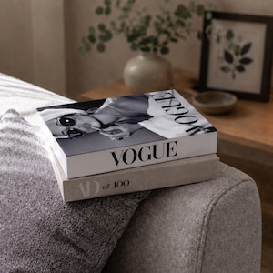 Könnte beinhalten: Ein Stapel aus zwei Hardcover-Büchern. Das obere Buch ist weiß mit dem schwarzen Schriftzug VOGUE und einem Schwarz-Weiß-Foto einer Person mit Sonnenbrille. Das untere Buch ist hellbeige mit dem schwarzen Aufdruck AD at 100 auf dem Buchrücken.