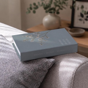 Puede incluir: Un libro decorativo rectangular de color azul con una ilustración de una rama floral blanca en la portada. El texto BREATHE BELIEVE RECEIVE está impreso en blanco tanto en la cubierta superior como en el lomo del libro, sobre una superficie gris.