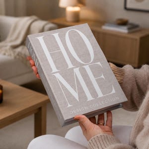 May include: A rectangular decorative book with a light beige fabric cover featuring the word HOME in large white capital letters and the text DESIGN COLLECTION printed in smaller white letters below. The book has a textured linen-like finish.