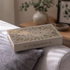 Puede incluir: Un libro rectangular con una cubierta de color crema que presenta un borde floral de pequeñas flores rosas y verdes. El centro de la cubierta muestra el texto "WHERE FLOWERS BLOOM SO DOES HOPE" dentro de un marco rectangular fino.