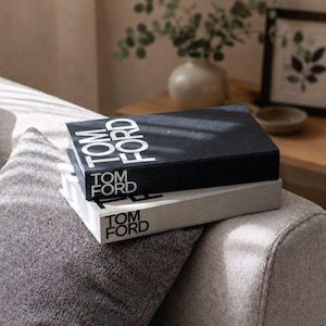 Könnte beinhalten: Zwei Hardcover-Bücher, die auf einer strukturierten Oberfläche gestapelt sind und jeweils den Schriftzug TOM FORD in fetten, weißen Großbuchstaben auf dem Buchrücken tragen. Das obere Buch hat einen schwarzen Einband mit genarbter Struktur.