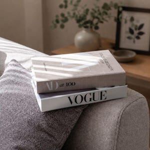 May include: Two stacked hardcover books resting on a textured grey surface. The top book is beige with the text AD at 100 A CENTURY OF STYLE. The bottom book is white with the word VOGUE printed in large black capital letters on the spine.