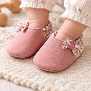 Könnte beinhalten: Ein Paar weiche Babyschuhe aus einfarbigem, altrosa Stoff mit strukturierter Oberfläche. Die Ferse und der Kragen sind mit einem cremefarbenen Blumenmuster mit kleinen rosa Blüten und grünen Blättern versehen. Jeder Schuh hat eine kleine passende Schleife.